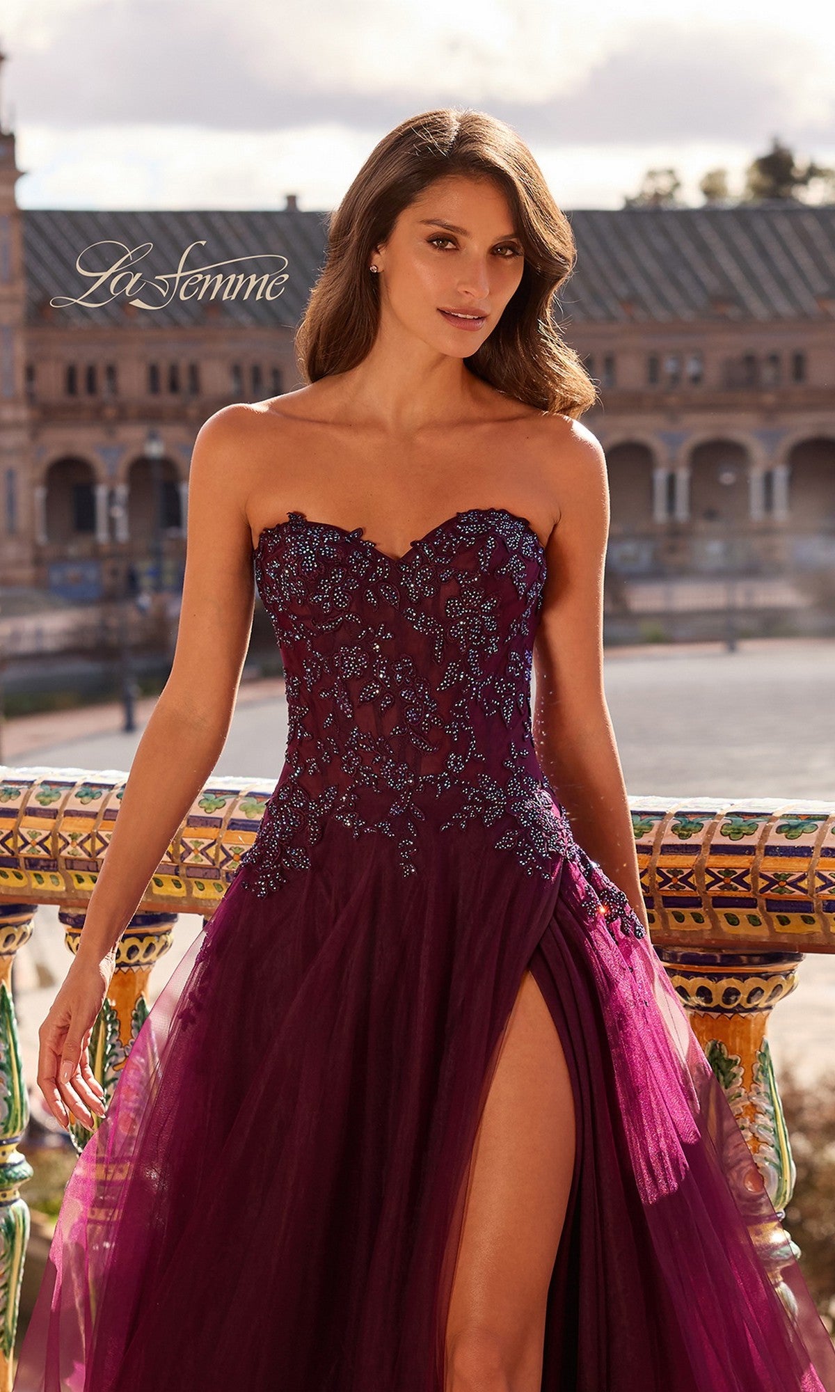 A woman wearing the La Femme 33551 long prom dress with a strapless corset bodice and dark purple details poses outdoors by ornate railings, with a grand historic building visible in the background.